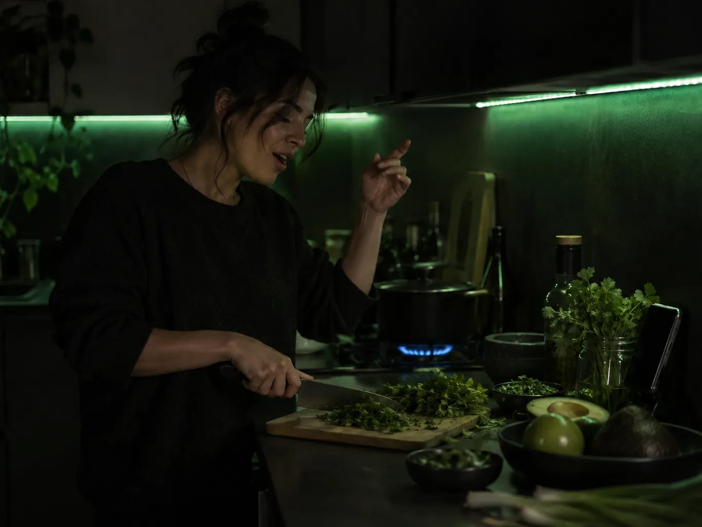Home cook chopping herbs at a dark kitchen counter lit by green under-cabinet lights, rapping along to music from a phone
