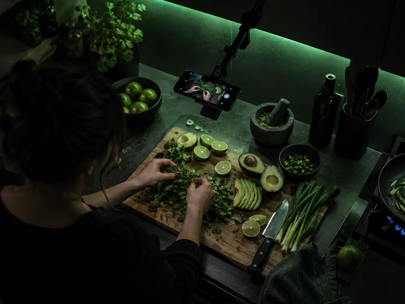 Overhead view of a food creator filming with a phone rig over a cutting board of sliced limes, avocado and herbs in a dark green-lit kitchen